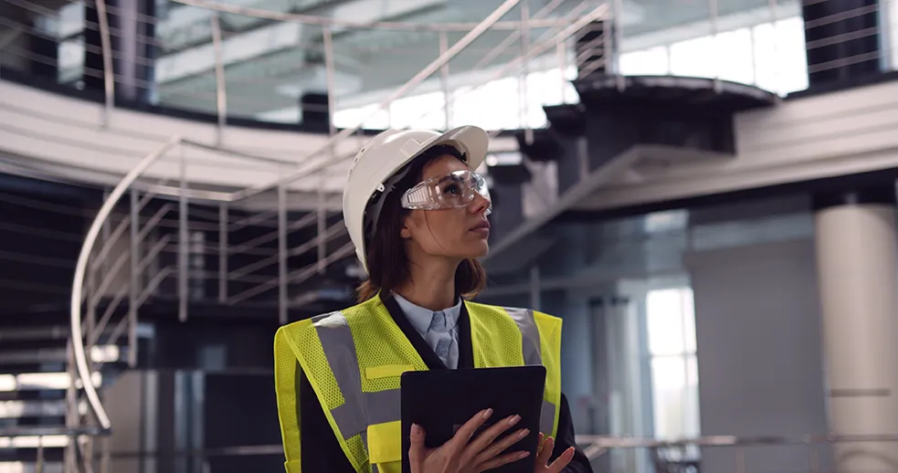 Women in real estate development: Female construction professional in safety gear reviewing building plans inside a modern commercial structure.